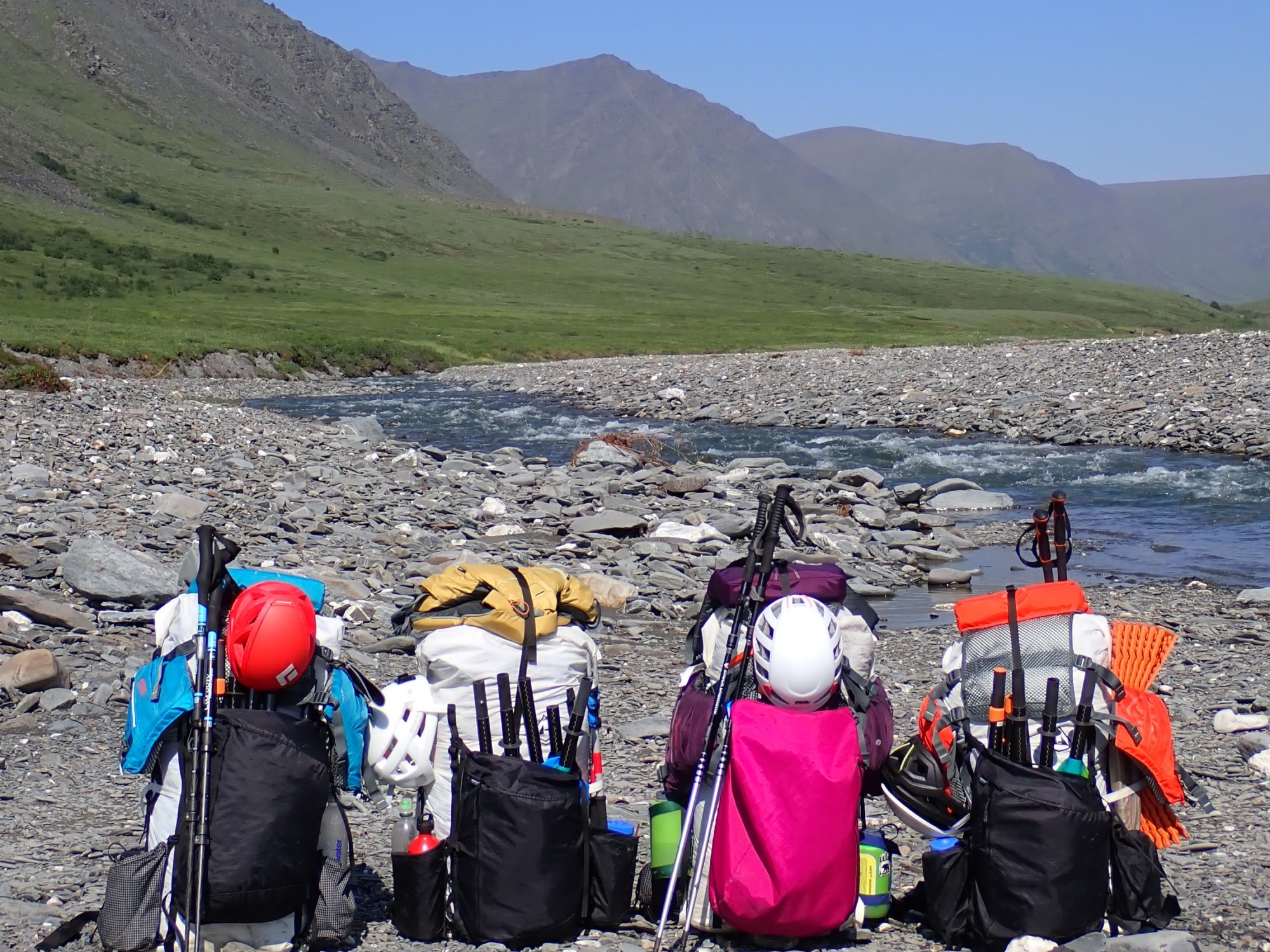 4 backpacks loaded with packrafting gear sit on a gravel bank of a river, mountains in background